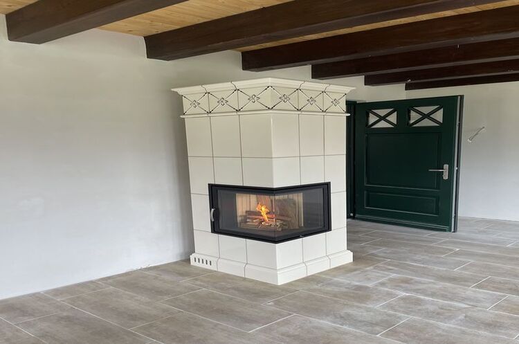 Moderner Kachelofen mit Feuer im hellen Wohnzimmer mit Holzbalkendecke und Fliesenboden.